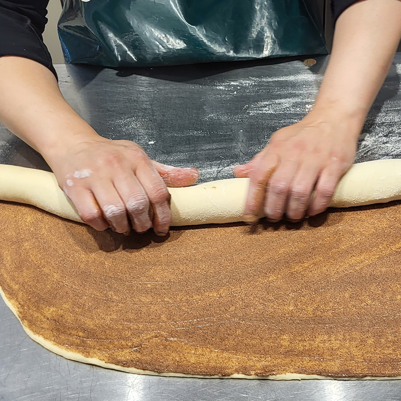 Zubereitung der Zimtschnecken: Der ausgerollte Teig wird händisch mit einer Butter-Zimt-Zucker-Mischung bestrichen und aufgerollt.