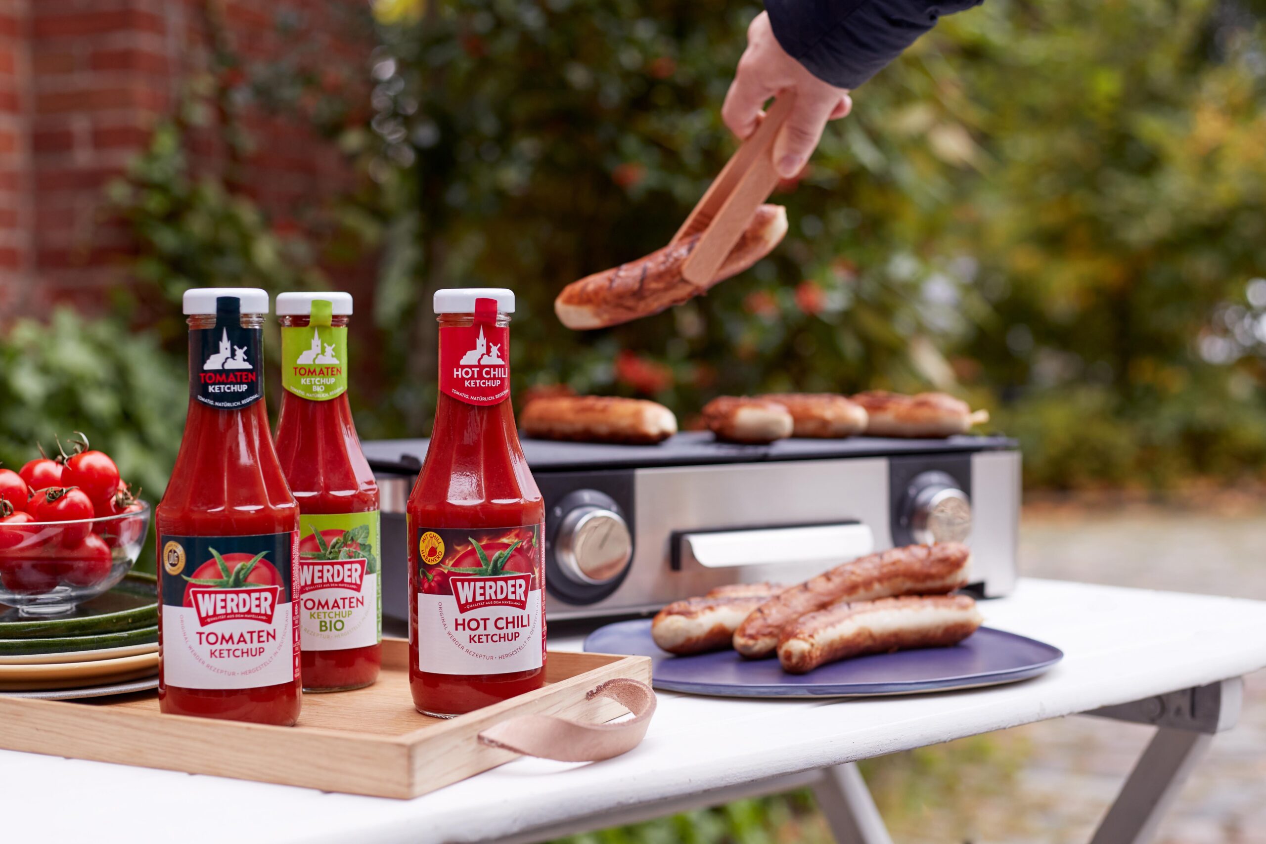 Drei Flaschen Werder Ketchup stehen vor einem Elektrogrill mit Bratwürsten.